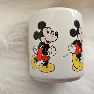 May include: White ceramic mug with a cartoon image of Mickey Mouse walking. The mug features two images of Mickey Mouse, one in the foreground and one in the background.