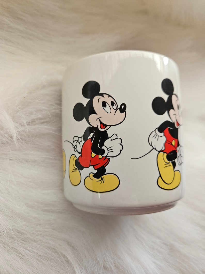 May include: White ceramic mug with a cartoon image of Mickey Mouse walking. The mug features two images of Mickey Mouse, one in the foreground and one in the background.