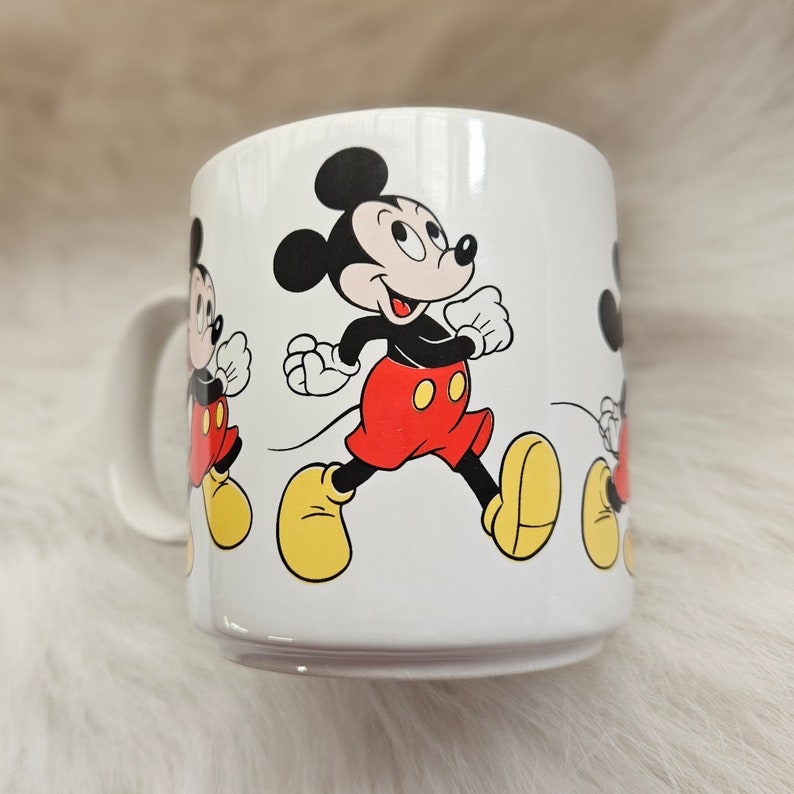 May include: White ceramic mug with a cartoon image of Mickey Mouse walking. The mug features three images of Mickey Mouse in different positions, all with a red and yellow color scheme.