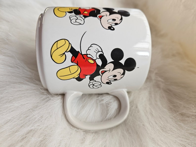 May include: White ceramic mug with a cartoon image of Mickey Mouse running. The mug has a handle and features two images of Mickey Mouse.