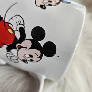 May include: White ceramic mug with a cartoon image of Mickey Mouse. Mickey is wearing red shorts and yellow shoes. He is running and smiling.