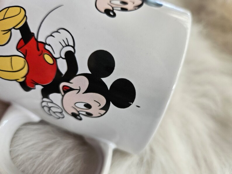 May include: White ceramic mug with a cartoon image of Mickey Mouse. Mickey is wearing red shorts and yellow shoes. He is running and smiling.