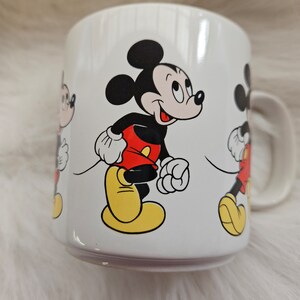 May include: White ceramic mug with a colorful cartoon image of Mickey Mouse. The mug features three Mickey Mouse characters walking in a line.