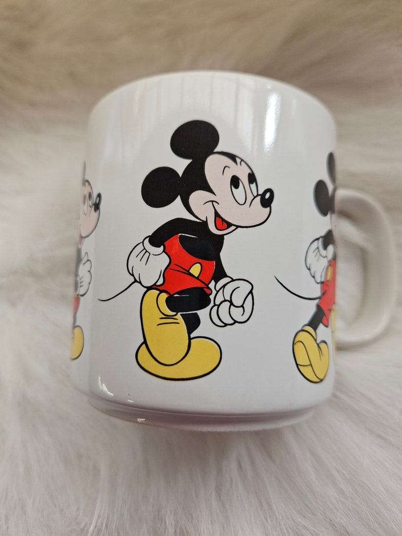 May include: White ceramic mug with a colorful cartoon image of Mickey Mouse. The mug features three Mickey Mouse characters walking in a line.