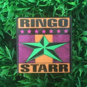 Puede incluir: Un pin rectangular con el nombre "RINGO STARR" en letras negras. El pin presenta una estrella verde sobre un fondo naranja, con estrellas rosas arriba y un patrón rosa ondulado.