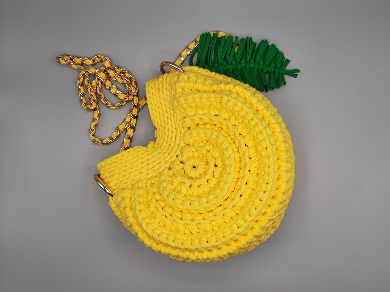 May include: A yellow crocheted handbag shaped like a lemon with a green crocheted leaf attached to the top. The bag has a gold chain strap.