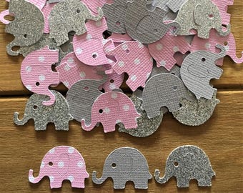 May include: A collection of paper elephant cutouts in pink, gray, and silver glitter. The pink elephants have white polka dots. The cutouts are scattered on a wooden surface, suitable for baby shower or party decorations.
