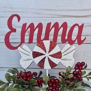 May include: A white cake with red berries and greenery. The cake is topped with a red and white candy cane and a red wooden sign that says "Emma".