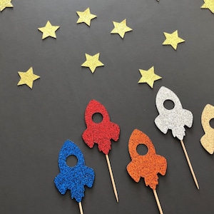 May include: Five glitter rocket ship cake toppers in different colors: red, blue, silver, orange, and gold. The toppers are on a black background with gold glitter stars.