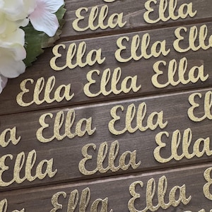 May include: Close-up of several wooden planks with the name "Ella" in gold glitter script. The name is repeated multiple times across the planks. White and pink flowers are in the upper left corner.