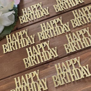 May include: Gold glitter "Happy Birthday" cake toppers arranged on a wooden surface. The toppers are in a cursive font and are evenly spaced. The background is a dark brown wood grain.