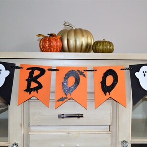 May include: A black and orange Halloween banner with the word "BOO" in black glitter letters and two white ghosts on either end. The banner is hanging on a white cabinet.