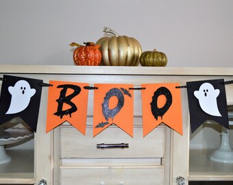 May include: A black and orange Halloween banner with the word "BOO" in black glitter letters and two white ghosts on either end. The banner is hanging on a white cabinet.