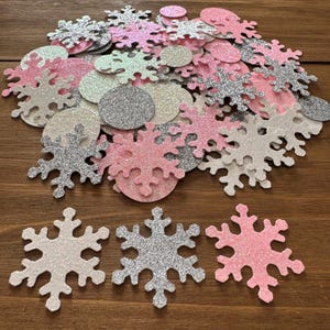 May include: A collection of glittery snowflake and circle confetti in shades of pink, silver, and white. The confetti is scattered on a wooden surface, creating a festive and decorative display. Ideal for winter-themed crafts and celebrations.