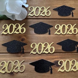 May include: Gold glitter "2026" cutouts and black glitter graduation caps are arranged on a wooden surface. The numbers and caps are scattered across the frame, suggesting a graduation theme. A white flower is in the upper left corner.