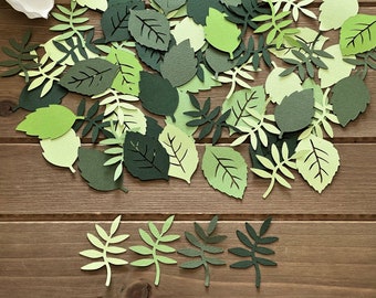 May include: A collection of green paper leaves of various shapes and sizes, scattered on a wooden surface. The leaves are cut out and have a delicate, intricate design.