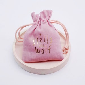 May include: A pink drawstring bag with a gold logo that says "stella and wolf" on it. The bag is sitting on a light pink round surface.