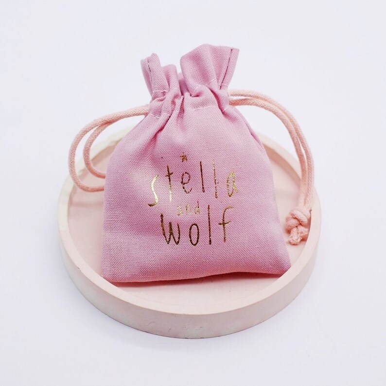 May include: A pink drawstring bag with a gold logo that says "stella and wolf" on it. The bag is sitting on a light pink round surface.