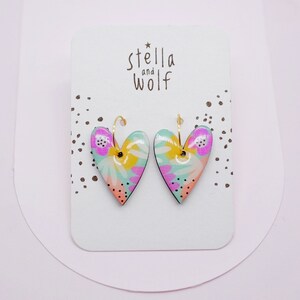 May include: A pair of heart-shaped earrings with a floral pattern in shades of pink, yellow, and green. The earrings are made of ceramic and have gold hoops.