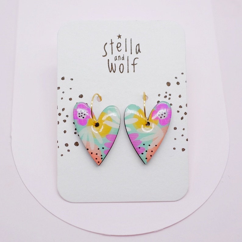 May include: A pair of heart-shaped earrings with a floral pattern in shades of pink, yellow, and green. The earrings are made of ceramic and have gold hoops.