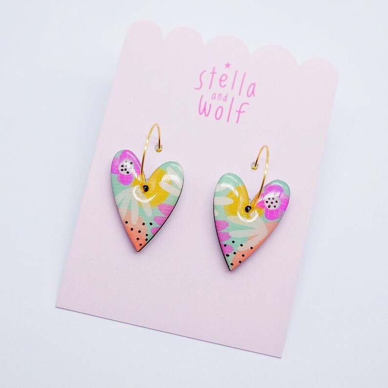 May include: A pair of gold hoop earrings with heart-shaped charms. The charms are decorated with a colorful floral pattern in shades of pink, yellow, green, and blue.