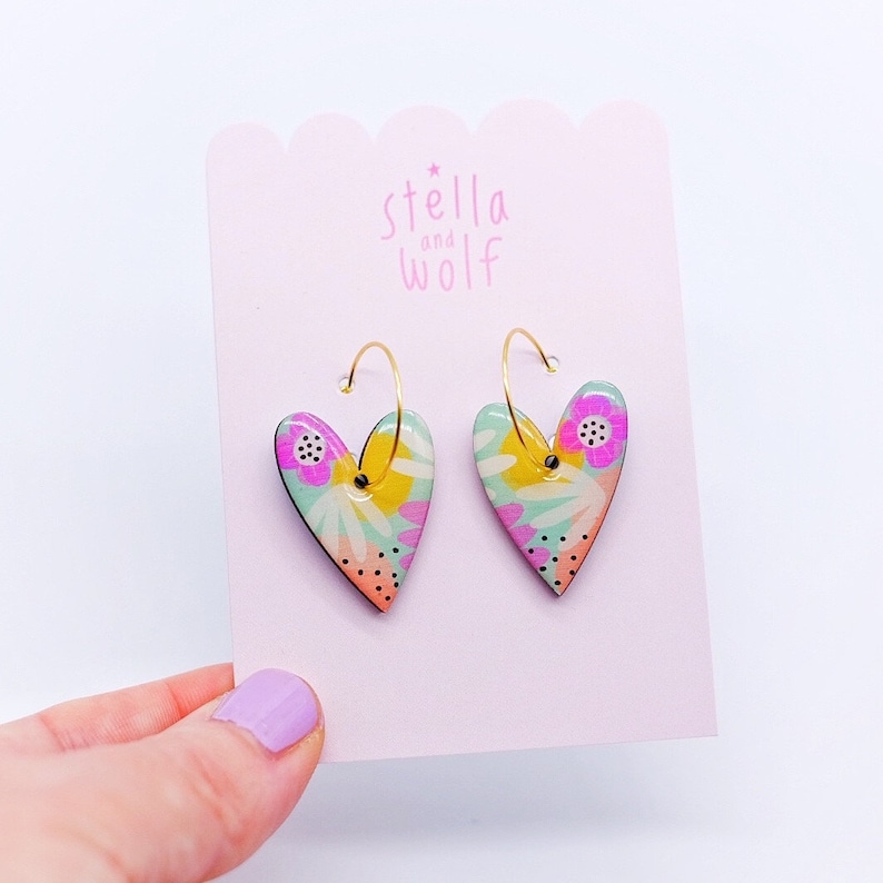 May include: A pair of gold hoop earrings with heart-shaped charms. The charms are decorated with a colorful floral pattern in shades of pink, yellow, green, and blue. The earrings are displayed on a pink card with the text "stella and wolf" printed on it.