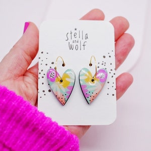 May include: A pair of gold hoop earrings with heart-shaped charms. The charms are decorated with a colorful floral pattern in shades of pink, yellow, green, and blue.