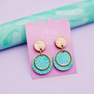 May include: A pair of turquoise blue and white polka dot earrings with gold accents. The earrings are made of wood and have a pink and gold resin top. The earrings are on a pink card with the text "Stella and Wolf" in white.