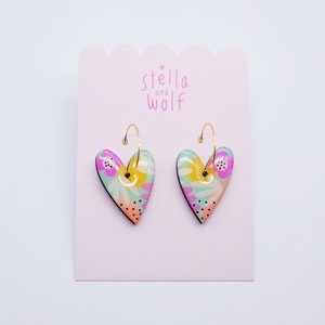 May include: A pair of gold hoop earrings with heart-shaped charms. The charms are decorated with a floral pattern in shades of pink, yellow, green, and orange.