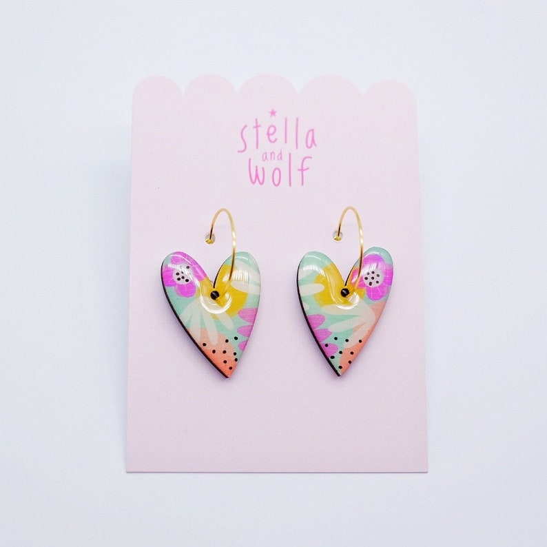May include: A pair of gold hoop earrings with heart-shaped charms. The charms are decorated with a floral pattern in shades of pink, yellow, green, and orange.