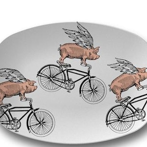 May include: A white oval ceramic serving platter with a repeating pattern of pink pigs with wings riding bicycles. The pigs are facing forward and the bicycles are black with white tires.