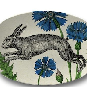 May include: An oval serving platter with a cream-colored background. The platter features a black and white illustration of a running hare amidst blue cornflowers and green foliage. A decorative piece for serving food.