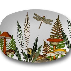 May include: An oval-shaped white plate featuring a detailed botanical illustration. The design includes various green and brown ferns, several orange and brown mushrooms of different sizes, a small brown bird, and a flying dragonfly.