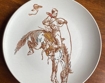 May include: A white ceramic plate with a brown and white illustration of a cowboy riding a bucking horse. The cowboy is wearing a hat and a red bandana. The horse is rearing up on its hind legs.