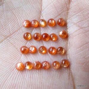 May include: A collection of 18 round, orange sunstone cabochons. The stones are arranged in rows on a hand, with each row containing six stones.