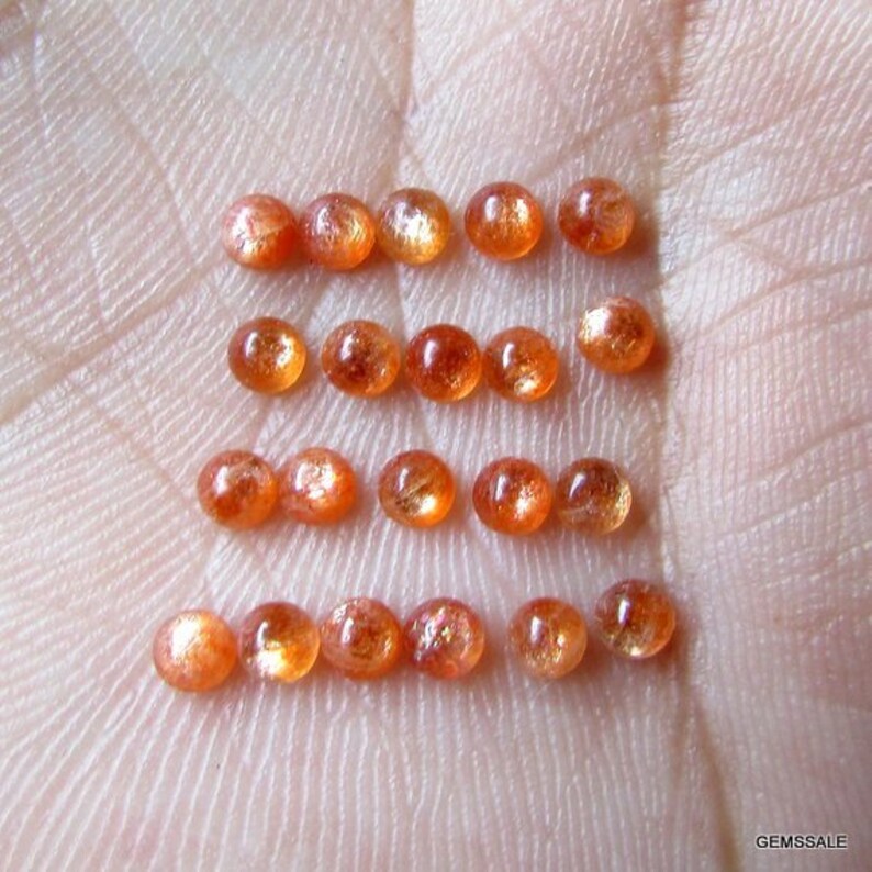 May include: A collection of 18 round, orange sunstone cabochons. The stones are arranged in rows on a hand, with each row containing six stones.