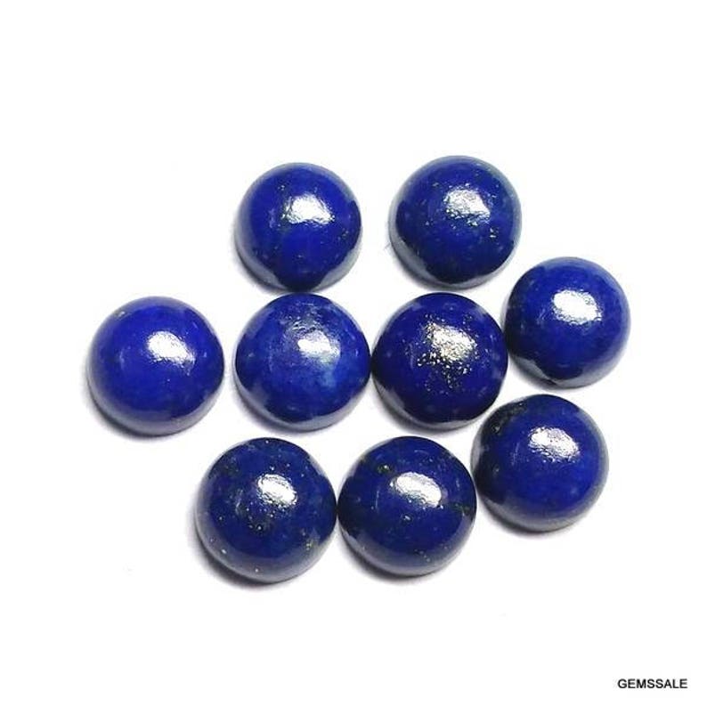 Pu&ograve; includere: Nove cabochon lucidati in lapislazzuli blu intenso, ciascuno con una forma arrotondata e bombata. Le pietre preziose sono disposte su uno sfondo bianco, mostrando il loro ricco colore e le inclusioni naturali. L'immagine &egrave; ben illuminata.