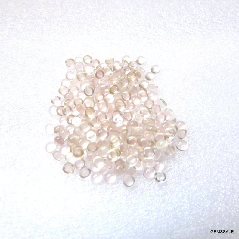 May include: A pile of translucent, pale pink, round gemstone cabochons. The cabochons are smooth and have a slightly iridescent sheen.