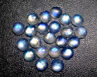 Puede incluir: Una colección de 20 cabujones de piedra lunar azul iridiscente. Las piedras son redondas y tienen una superficie lisa y pulida. Están dispuestas en un montón suelto sobre un fondo negro.