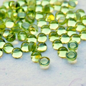 Puede incluir: Una vista de cerca de una gran cantidad de pequeñas piedras preciosas de peridoto redondas y verdes. Las piedras preciosas son lisas y brillantes, y están dispersas sobre una superficie blanca.
