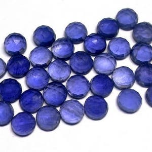 May include: A group of 25 faceted round blue gemstones, each approximately 6mm in diameter. The gemstones are arranged in a loose pile on a white surface.