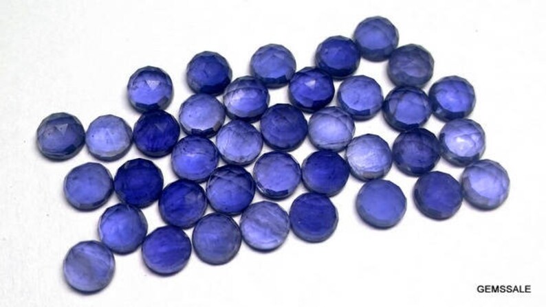 May include: A group of 25 faceted round blue gemstones, each approximately 6mm in diameter. The gemstones are arranged in a loose pile on a white surface.