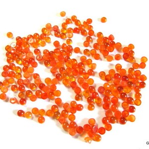 May include: A large group of small, round, orange carnelian cabochons. The cabochons are scattered on a white surface.