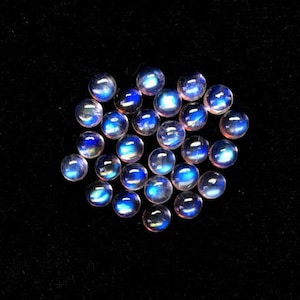 Puede incluir: Un grupo de 18 cabujones de piedra de luna azul iridiscente, cada uno con una superficie lisa y redondeada. Las piedras est&aacute;n dispuestas en un grupo suelto sobre un fondo negro.