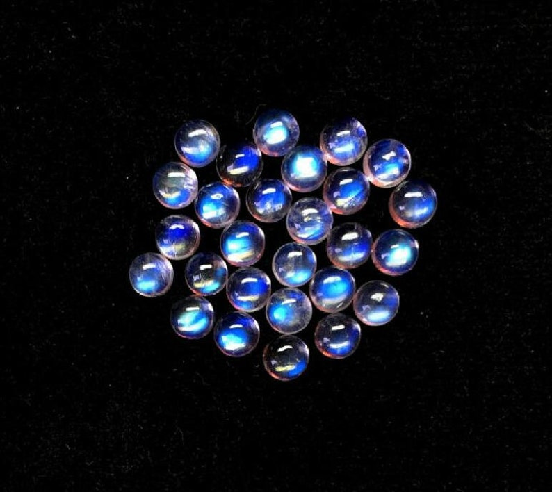 Puede incluir: Un grupo de 18 cabujones de piedra de luna azul iridiscente, cada uno con una superficie lisa y redondeada. Las piedras est&aacute;n dispuestas en un grupo suelto sobre un fondo negro.