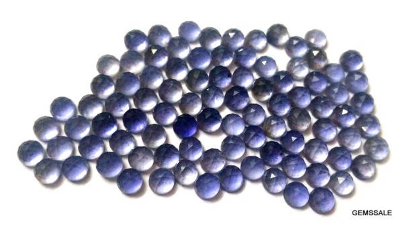 May include: A collection of round, faceted, purple gemstones. The gemstones are arranged in a loose pile and have a variety of shades of purple.
