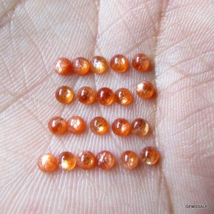 May include: A collection of 18 oval-shaped sunstone gemstones, ranging in color from orange to reddish-brown. The stones are arranged in three rows of six, with each stone reflecting light.