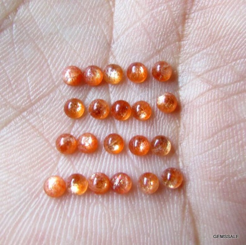 May include: A collection of 18 oval-shaped sunstone gemstones, ranging in color from orange to reddish-brown. The stones are arranged in three rows of six, with each stone reflecting light.