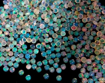 May include: A scattering of small, round opals. The opals are translucent and show a variety of colours, including white, blue, green, and red, against a dark background. These gemstones are likely for jewellery making.