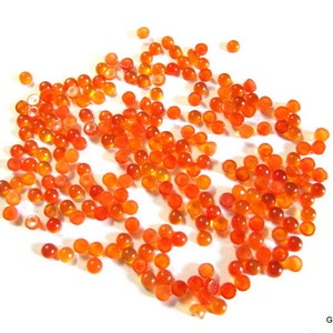 May include: A large group of small, round, orange carnelian cabochons. The cabochons are scattered on a white surface.